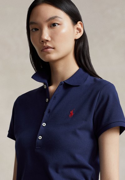 Ung kvinne med rett svart hår iført en marineblå polotrøye med en rød brodert logo, stående mot en enkel lys bakgrunn.