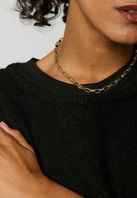 Collier en chaîne dorée avec des maillons rectangulaires, porté sur un pull noir texturé. Vue en gros plan de l'encolure et de la zone des épaules.