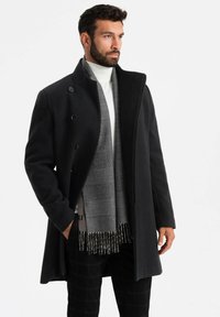 Abrigo de mezcla de lana negro con cuello alto y botones de doble botonadura, combinado con una bufanda de cuadros grises con bordes deshilachados.