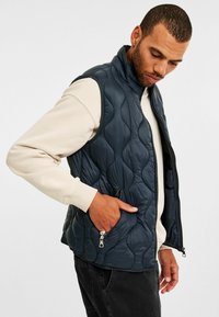 Man med marinblå kviltad ärmlös väst över beige sweatshirt, med hand i västfickan, blickande ner mot vit bakgrund.