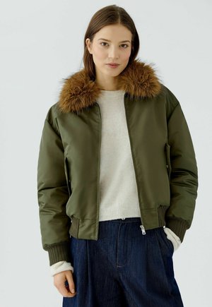 Vrouw draagt groene bomberjas met bruine bontkraag, witte trui en blauwe broek, staand tegen een effen lichte achtergrond.