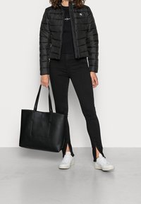 Svart puffjacka med dragkedja fram och broderad logotyp, kombinerad med svarta skinny jeans med sidopenningar och vita sneakers. Svart axelväska.