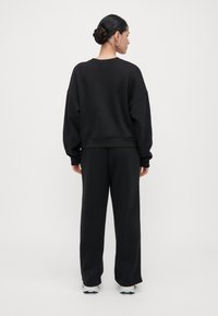 Mulher com coque trançado, vestindo um sweatshirt preto e calças largas pretas, de pé de costas em um fundo branco liso.