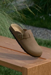 Braune Wildleder-Slipper mit verstellbarem Schnallenriemen, die auf einer Holzbank im Freien liegen, mit grünem Gras und Pflanzen im Hintergrund.