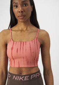 Sieviete, tērpusies rozā krāsā krāsotā topā ar plānām siksnām un brūnās Nike Pro leggingas, stāv pretī vienkāršam gaišam fona.