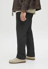 Black trousers made of a smooth fabric, straight-leg cut, worn with beige suede shoes. Brown leather jacket with a zipper visible on the side.