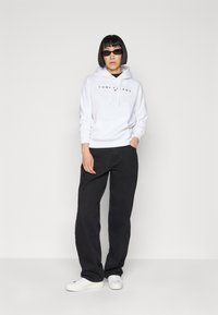 Felpa bianca con logo "TOMMY JEANS" in nero, abbinata a pantaloni neri larghi e sneakers bianche. Il modello indossa occhiali da sole sottili.