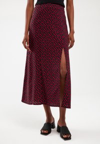 Red and black midi skirt with a geometric heart pattern, featuring a side slit, made of smooth fabric; paired with black textured slide sandals.
