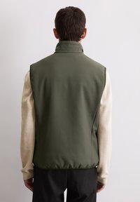 Ciemnozielona, bezrękawnik z wysokim kołnierzem, wykonany z gładkiej tkaniny. Posiada dopasowany fason i jest noszony na jasnozabarwionej, teksturowanej, długorękawowej bluzce.