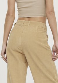 Pantalon en velours côtelé beige clair avec des côtes verticales, présentant une taille ajustée et une coupe décontractée le long des jambes, vu de l'arrière.