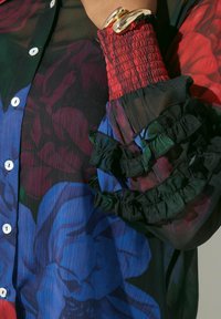 Blusa floreale trasparente con motivi blu, rossi e verdi. Caratterizzata da polsini arricciati neri e rossi, chiusura con bottoni e tessuto testurizzato.