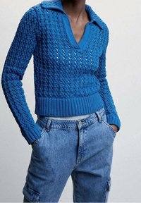 Blauer Strickpullover mit V-Ausschnitt und Kragen, mit strukturiertem Muster. Kombiniert mit hellblauen Denim-Cargohosen.