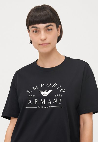Melna kokvilnas T-krekls ar baltu Emporio Armani logo un ērgļa emblēmu centrā priekšpusē. Īsas piedurknes un apaļais kakls.