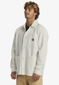 Light-colored button-up jacket in soft fabric, featuring a chest pocket, two front pockets, and a relaxed fit design with a collar.