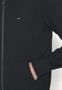 Sudadera con cremallera en color azul marino, confeccionada en una mezcla de algodón, con un pequeño logo bordado en el pecho, puños de canalé y bolsillos laterales.