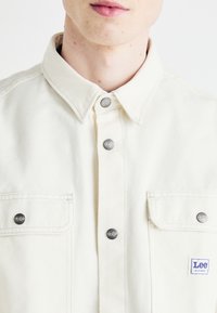 Lee skjorta med knapp, i off-white tyg, med metallknappar med logotyp, två bröstfickor och kontrasterande sömnadsdetaljer.