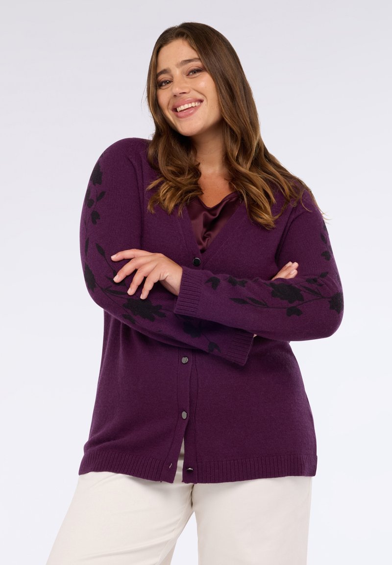 Leende kvinna med långt brunt hår, klädd i en lila kofta med svarta blommönster och vita byxor, med armarna korsade självförtroende.