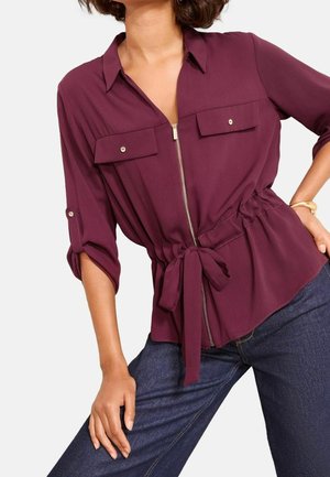 Mujer que lleva una blusa burdeos con botones, bolsillos delanteros, cremallera y cinturón anudado a la cintura, combinada con vaqueros azul oscuro.