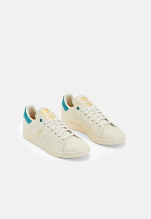 Par off-white lædersneakers med grønblå hælkapper, hvide snørebånd og guldfarvede logodetaljer på tunge og side, vist på hvid baggrund.