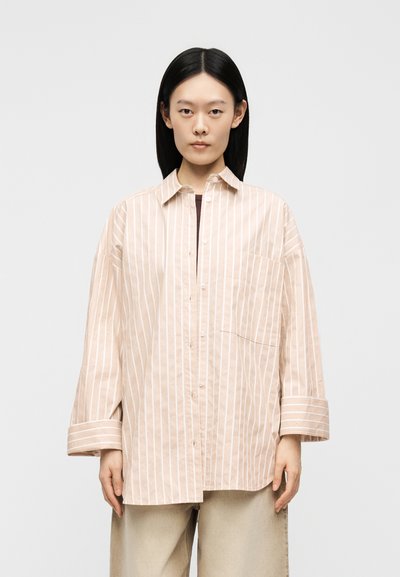 Femme aux cheveux noirs et raides, portant une chemise beige oversize avec des rayures verticales blanches et un pantalon beige, debout devant un fond uni.