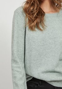 Svetle zelený svetr vyrobený z měkké tkaniny, s volným střihem, kulatým výstřihem a dlouhými rukávy. Černé džíny viditelné v pase.