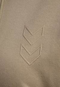 Beżowy materiał z teksturowaną powierzchnią, z dwoma wzniesionymi kształtami chevron w pasującym kolorze, umieszczonymi centralnie.