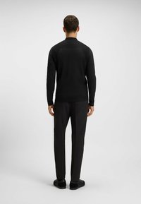 Schwarzer Pullover mit langen Ärmeln und hohem Kragen, mit einem strukturierten Rückenteil und klaren Linien, kombiniert mit dunklen Hosen und schwarzen Sneakers.