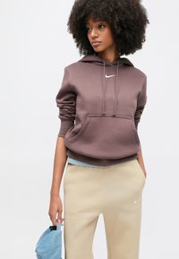 Barna kapucnis pulóver húzózsinórokkal és első zsebbel, kis fehér logóval díszítve. Bézs nadrággal párosítva, kezében világoskék sapkával.