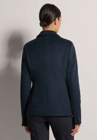 Marineblauer Wollblazer mit taillierter Passform, Reverskragen und geknöpften Manschetten. Glatte Textur, strukturierter Schnitt, mit Nahtdetails auf der Rückseite.