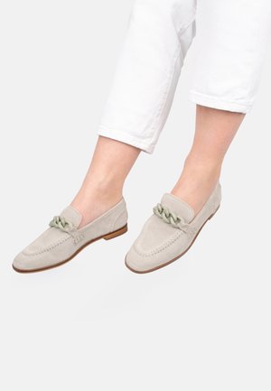 Hellbeige Wildleder-Loafer mit klobiger Kettendetail, getragen zu weißen, verkürzten Hosen, auf Füßen vor einfarbigem Hintergrund gezeigt.