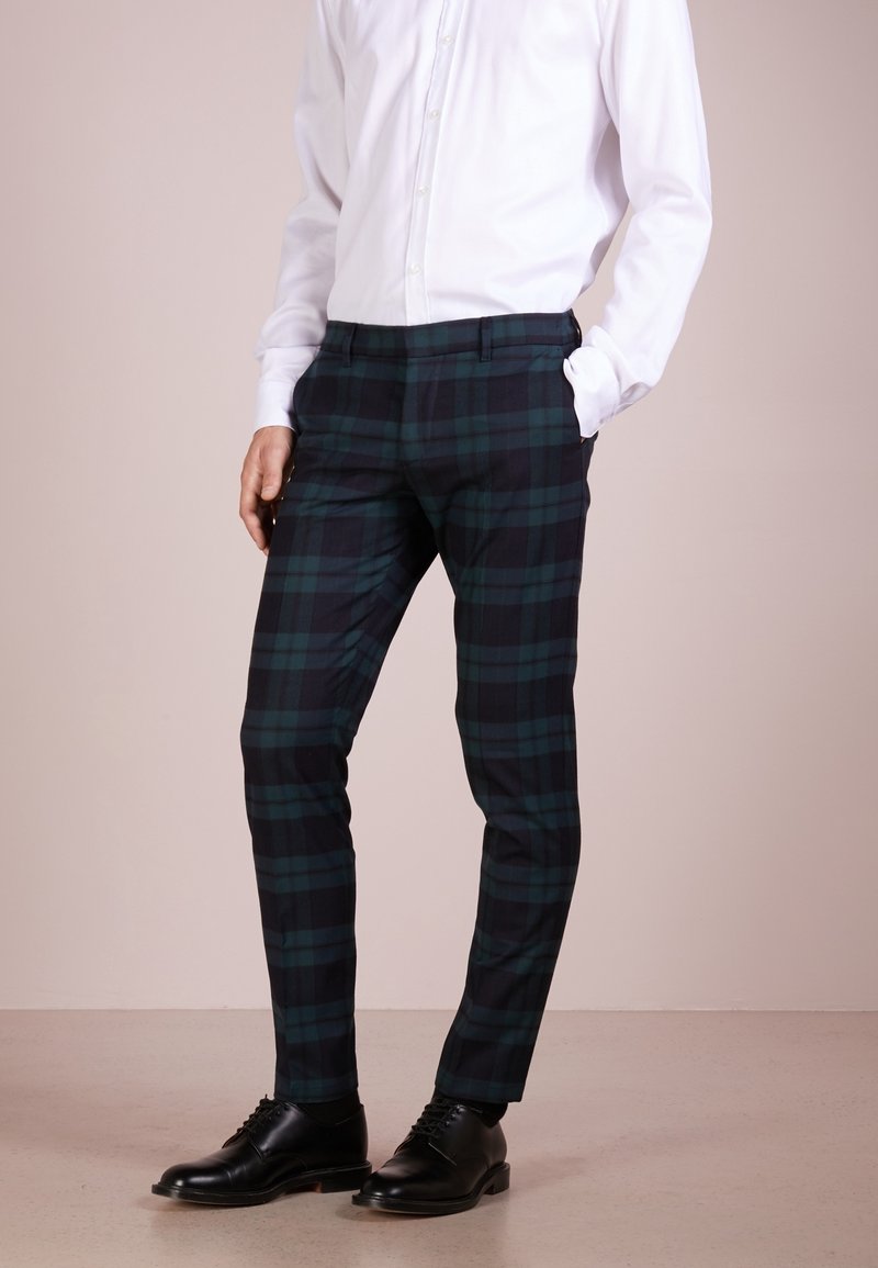 Pantalones con un patrón a cuadros en azul marino oscuro y verde, de corte entallado, con frente plano y bolsillos laterales, combinados con zapatos negros y una camisa blanca.