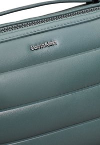 Borsa a tracolla in finta pelle color teal con cuciture diagonali, zip in argento e logo Calvin Klein. Texture liscia con un design strutturato.