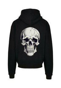 Černá mikina s kapucí s velkým, podrobným bílým potiskem lebky na zádech, vybaveným jemným stínováním a prasklinami pro texturu.