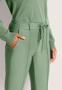 Vrouw in lichtgroene hoge taillebroek met naden aan de voorkant en een trekkoord, hand in de zak, gecombineerd met een bijpassend groene langemouwen top.