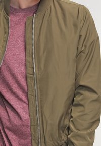 Olivově zelená bomber bunda se zipem vpředu, pružnými manžetami a lemem, nošená přes vínové tričko s kulatým výstřihem. Hladká textura, volný střih.