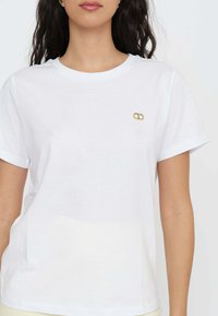 T-shirt bianco in cotone con maniche corte, scollo rotondo e un piccolo logo dorato sul petto. Tessuto morbido, vestibilità rilassata.