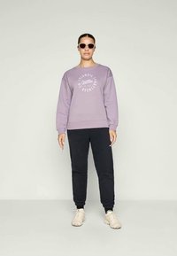 Sweatshirt lilás com logotipo branco, emparelhado com calças de jogging pretas. O modelo usa óculos de sol e ténis de cor clara. Design minimalista.