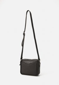 Bolso cruzado de piel negra con cierre de cremallera, forma rectangular, superficie texturizada y correa ajustable. Presenta un logo sutil en la parte frontal.