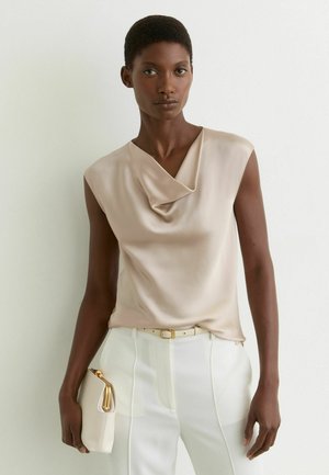 Femme aux cheveux courts portant un top en satin beige sans manches, un pantalon blanc et tenant une petite pochette beige sur fond blanc.