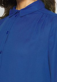 Chemise bleu à col classique, en tissu lisse, avec des boutons bleus assortis le long de la patte de boutonnage. Rassemblements subtils aux épaules.