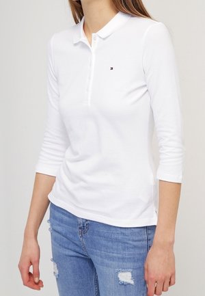 Kobieta ubrana w biały, długi rękaw polo z małym logo na piersi oraz podniszczone niebieskie dżinsy.