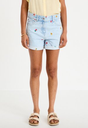 Kvinde iført lyseblå denimshorts med små farverige blomsterbroderier, en gul ærmeløs skjorte og beige sandaler med spænde.