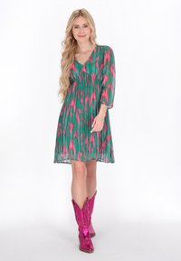 Rochie cu un model verde și roz, cu mâneci trei sferturi și decolteu în V, purtată cu cizme cowboy lucioase roz.