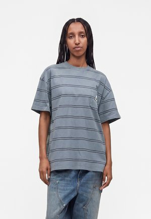 Jeune personne aux cheveux tressés portant un t-shirt ample gris rayé et un jean bleu délavé, debout devant un fond clair uni.