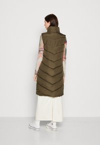 Gilet olive vert rembourré avec un col montant et un motif en chevrons ; porté par-dessus une longue jupe blanche et des chaussures Converse.
