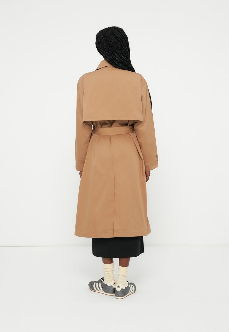 Tan trenchcoat med dobbeltknap design, bælte i taljen og rygkap detalje, lavet af glat stof, båret over en sort nederdel.