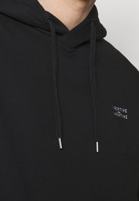 Černá mikina s kapucí vyrobená z měkké tkaniny, má klokaní kapsu, šňůrky s metalickými konci a vyšívané logo na hrudi.