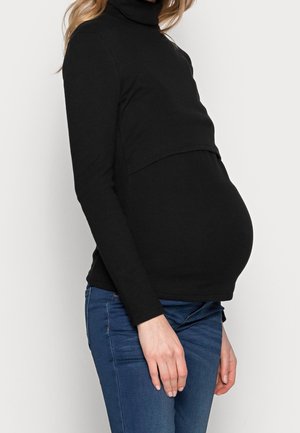 Zwangere vrouw die een zwart coltrui met lange mouwen en een blauwe spijkerbroek draagt, zijdelings weergegeven tegen een effen achtergrond.