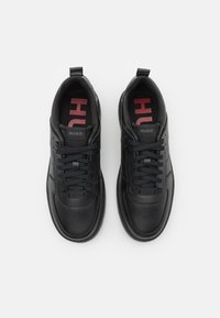 Sneakers in pelle nera con una texture liscia, punta perforata, design con lacci e logo "HUGO" rosso a contrasto sulle solette.