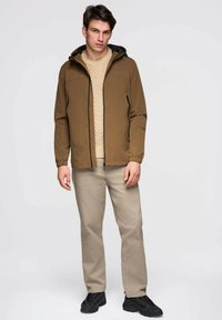Veste à capuche marron avec des accents noirs, fermeture éclair, et deux poches avant, superposée à un pull en tricot crème et un pantalon beige.